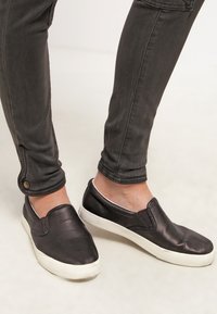 Personne portant un pantalon gris foncé ajusté et des baskets noires sans lacets avec des semelles blanches, debout sur un sol clair.