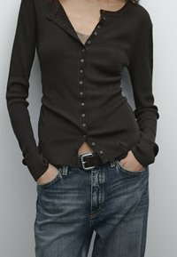 Donkerbruine geribbelde langemouwtop met een knoopsluiting aan de voorkant en manchetten, gecombineerd met lichtblauwe denimjeans en een zwarte riem met een zilveren gesp.