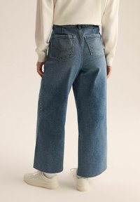 Szerokie, niebieskie jeansy z surowym wykończeniem i dwiema tylnymi kieszeniami. Noszone z białymi sneakersami, ukazującas casualowy styl i jasny odcień.