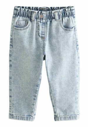 Enfant portant un jean taille élastique bleu clair, une chemise crème à manches courtes et des chaussures beiges à enfiler avec des taches noires, debout les mains dans les poches.