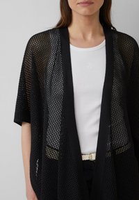 Zwarte gebreide cardigan met een open mesh ontwerp, contrasterende solide randen, gedragen over een witte ribbel top, gecombineerd met een geweven crème riem.