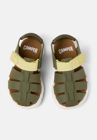 Grön sandal med ett texturerat ovandel som har korsande remmar. En mjuk brun innersula och vit gummisula, kompletterad med ett justerbart gult band.