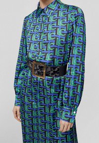 Skjorta med krage i ett geometriskt mönster i grönt, blått och svart; har en åtdragen midja med ett svart bälte och guldbucklad dekor.