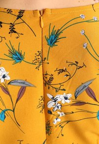 Keltainen kangas, jossa on valkoisia, sinisiä ja mustia kukkakuoseja. Kuvio sisältää erilaisia kasvien yksityiskohtia ja takana vetoketjukiinnityksen.