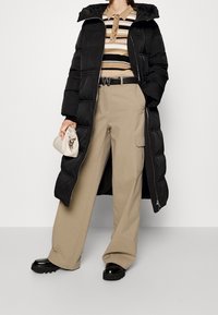 Schwarze Pufferjacke mit Kapuze, gestreiftes Hemd mit Kragen, beigefarbene Cargo-Hose und schwarze Plateauschuhe. Hält eine kleine beigefarbene Clutch.