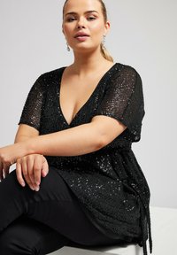 Top nero con paillettes, maniche corte e scollatura a V profonda. Presenta una vita aderente con un dettaglio di nodo. Indossato sopra pantaloni neri.