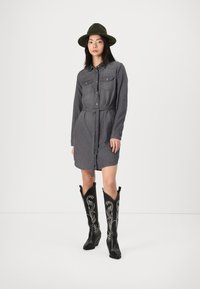 Robe chemise en denim gris avec deux poches poitrine et une taille ceinturée, associée à des bottes de cow-boy noires avec des coutures blanches décoratives.