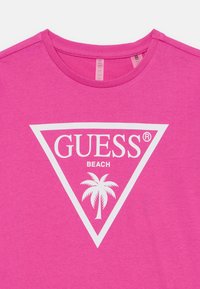 Roosa puuvillane t-särk, millel on valge kolmnurkne graafika, mille keskel on tekst "GUESS BEACH" ja palmipuu muster.