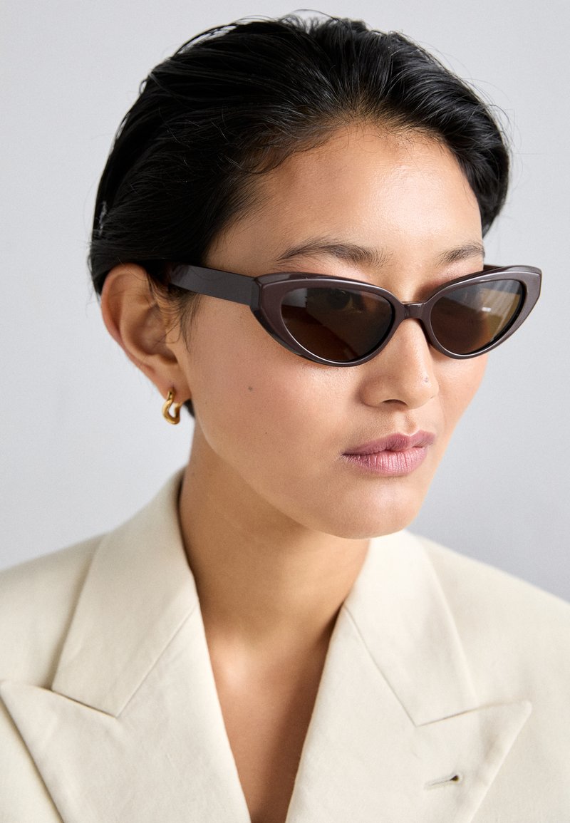 Barna macskaformájú napszemüveg sötét lencsékkel, vastag kerettel és sima felülettel, bézs blézerrel és arany fülbevalóval párosítva.