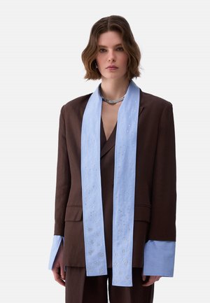 Giovane donna con capelli corti castani che indossa un completo marrone oversize e una lunga sciarpa azzurro chiaro con piccole decorazioni argento.