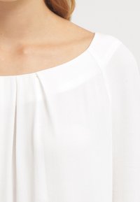 Weiße Bluse mit weitem Ausschnitt, weichem Material und dezenten Faltendetails in der Schulterpartie.