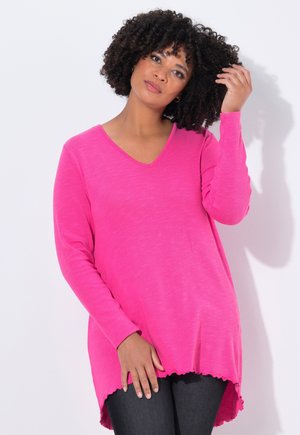 Un haut long à manches roses vives avec un col en V, présentant un ourlet festonné et une texture douce, associé à un jean foncé.