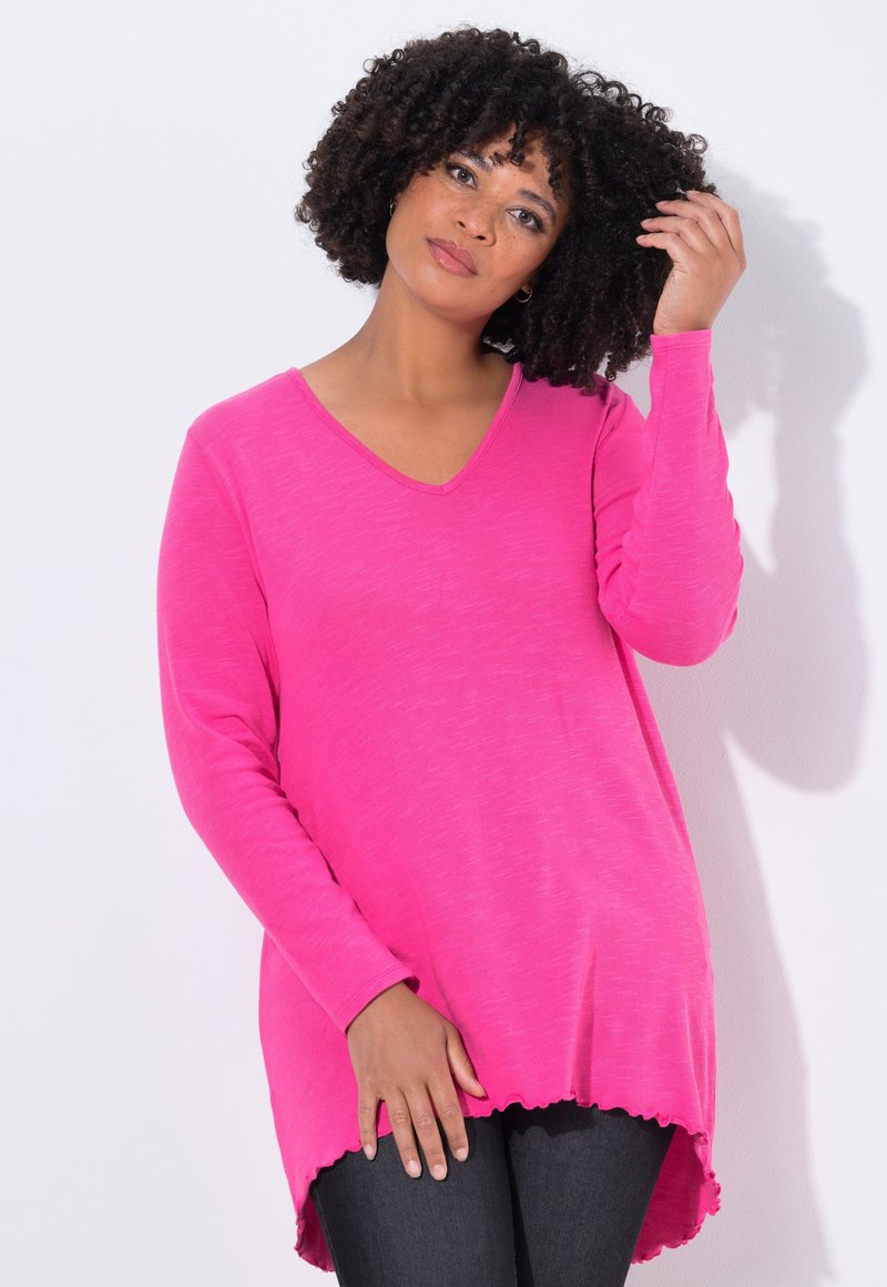 Maglietta lunga a maniche rosa vivace con scollatura a V, caratterizzata da un orlo ondulato e una texture morbida, abbinata a jeans scuri.