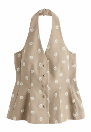 Kvinde med mørkt hår i en beige mønstret halterneck-top og shorts justerer sit hår mod en hvid væg og sten gulv baggrund.