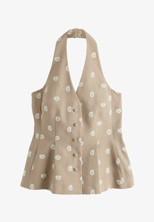 Sötét hajú nő bézs mintás nyakpántos felsőben és rövidnadrágban haját igazítja fehér fal és kőpadló előtt.