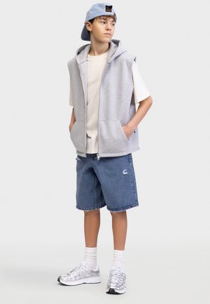 Αγόρι που φοράει μπλε καπέλο προς τα πίσω, γκρι αμάνικη μπλούζα με κουκούλα, λευκό μπλουζάκι, τζιν σορτς, λευκές κάλτσες και ασημένια αθλητικά παπούτσια, στέκεται με τα χέρια στις τσέπες.