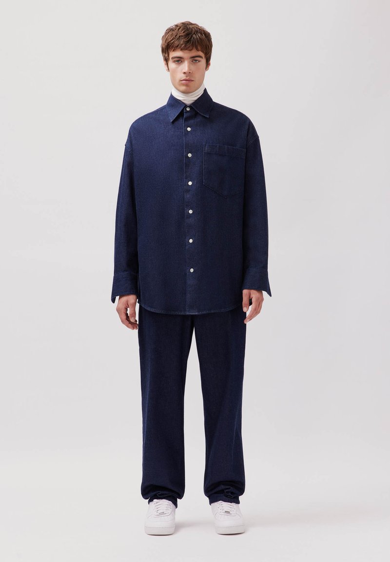 Marineblaue Jeanshemd und passende Hose, mit einer Tasche, Knöpfen und einem bequemen Schnitt. Kombiniert mit weißen Sneakers und einem hochgeschlossenen Oberteil.