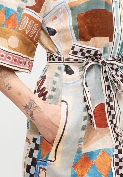 Χέρι ατόμου με τατουάζ στην τσέπη πολύχρωμου φορέματος που διαθέτει γεωμετρικά και αφηρημένα μοτίβα με ζώνη καρό δεμένη στη μέση.