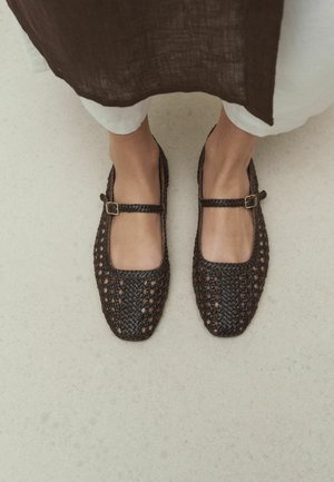 Feet wearing dark brown woven Mary Jane shoes with buckle straps, standing on a light floor, with part of white pants and brown garment visible.