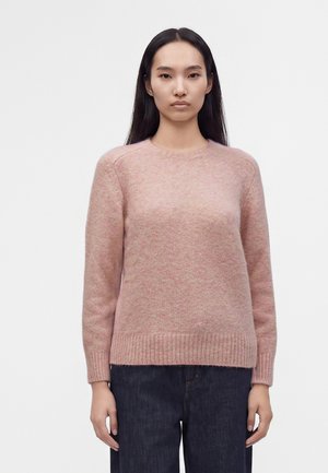 Femme aux longs cheveux noirs portant un pull en maille rose clair et un jean bleu foncé, debout devant un fond blanc uni.