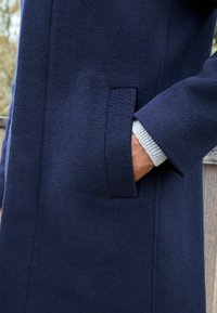 Cappotto blu navy realizzato in lana texturizzata, con una tasca laterale e un polsino in maglia grigia chiara a contrasto visibile sotto la manica.
