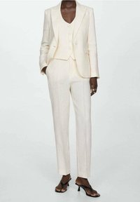 Traje de tres piezas en tejido beige claro, que incluye una blazer ajustada, un chaleco con botones y unos pantalones ajustados, combinados con tacones negros con tiras.