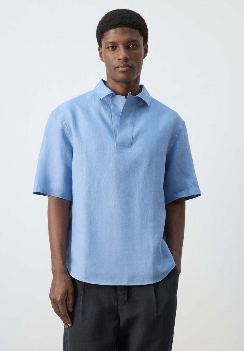 Jeune homme aux cheveux courts portant une chemise bleu clair à manches courtes avec col et un pantalon foncé sur un fond blanc uni.