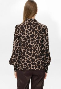 Donna con capelli lunghi che indossa una blusa a stampa animale marrone e nera con maniche a sbuffo, in piedi di spalle su uno sfondo semplice.