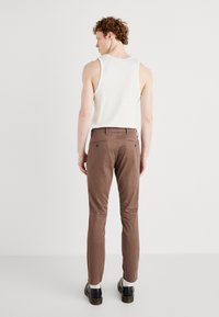 Brown cotton trousers with a slim fit, featuring back pockets and a clean design. Paired with a white sleeveless top and black shoes.