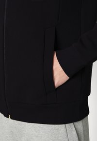 Svart sweatshirt med dragkedja, ribbade muddar, sidofickor och en slät yta. Matchas med ljusgrå byxor.