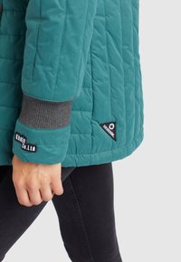 Personne portant une veste matelassée bleu sarcelle avec les logos visibles "Khujo Co. Ltd" et "Khujo Genuine", associée à un pantalon noir.