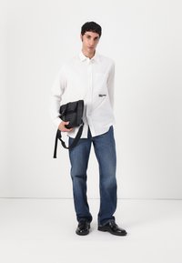 White button-up shirt with a front pocket, black messenger bag with adjustable strap, blue distressed jeans, and black shoes. Simple design.