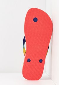 Sandale tong à semelle rouge avec connecteurs de sangles bleues et sangles latérales multicolores visibles, positionnée verticalement contre un coin blanc.