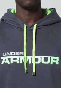 Sudadera gris con un interior verde, que presenta un gran logo en blanco y verde de "UNDER ARMOUR" y cordones de contraste en verde neón.