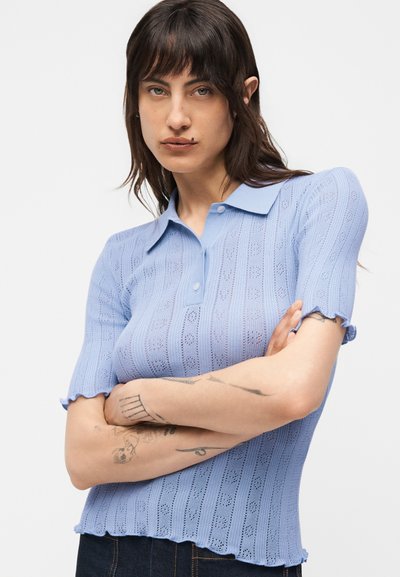 Γυναίκα με σκούρα μαλλιά που φοράει ανοιχτό μπλε πλεκτό πόλο με κοντά μανίκια, με σταυρωμένα τα χέρια, δείχνοντας τατουάζ στα αντιβράχια σε ένα απλό φόντο.