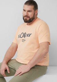 Lichtoranje t-shirt met marineblauw gedrukt logo, ronde hals, korte mouwen. Gecombineerd met olijfgroene broek. Effen achtergrond.