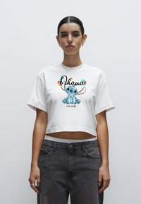 Biały krótki t-shirt z kolorową grafiką Stitche'a oraz napisem "Ohana oznacza rodzinę", zestawiony z szarymi, wysokim stanie jeansami.