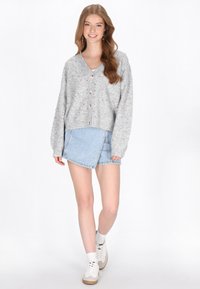 Fiatal nő sétál, szürke gombos kardigánt visel színes pöttyökkel, világoskék aszimmetrikus farmernadrágot, fehér zoknit és fehér sportcipőt.