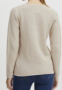 Pull beige en tricot avec un motif texturé, encolure ronde, manches longues et ourlet droit, porté avec un jean bleu.