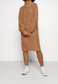 Abito in maglia a coste marrone con collo alto, maniche lunghe, taglio al ginocchio, abbinato a sneaker alte bianche.