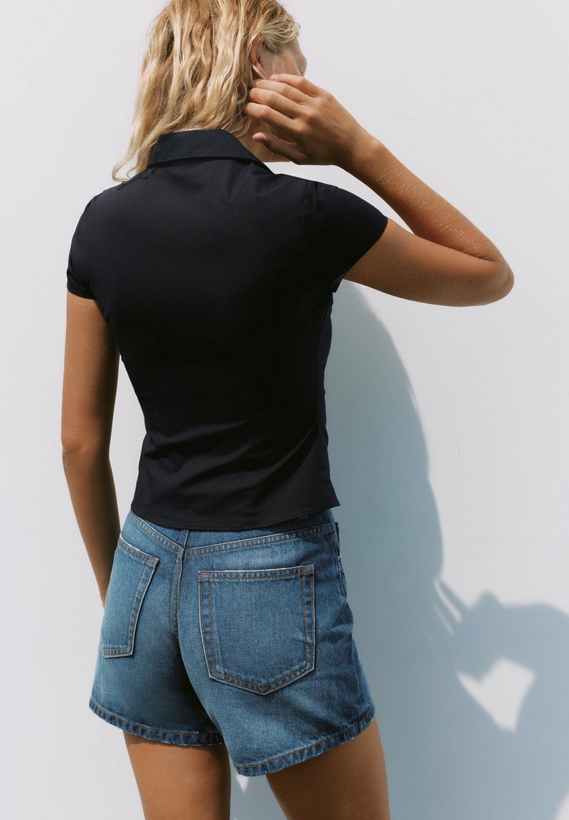 Femme aux cheveux blonds portant un t-shirt noir à manches courtes et un short en jean, debout contre un mur blanc, de dos, touchant sa tête.