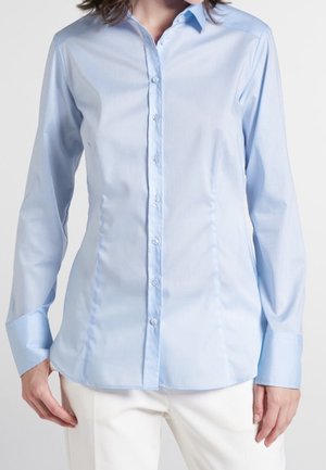 Chemise bleu clair à manches longues avec des boutons, portée par une personne en pantalon blanc, montrée des épaules jusqu'aux hanches sur un fond uni.