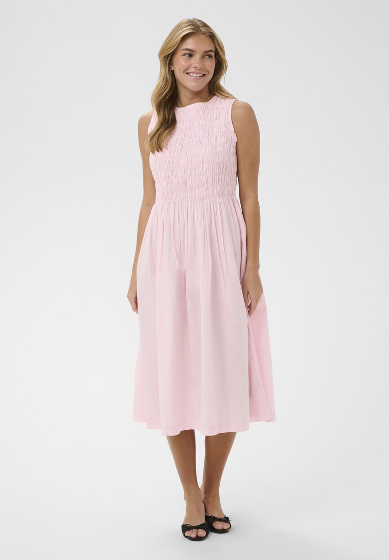 Femme en robe midi sans manches rose pâle avec corsage smocké, debout pieds nus sur fond blanc, souriante et regardant à droite.