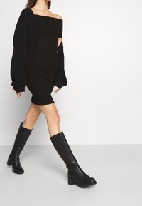 Abito nero a coste con scollo off-shoulder e arricciature, abbinato a stivali alti neri chunky. L'abito ha una silhouette aderente e un orlo sfrangiato.