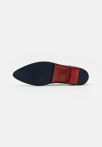Semelle en cuir marron avec un fond en caoutchouc noir, présentant une pointe effilée et un accent en cuir rouge près du talon. Détail de logo gravé visible.