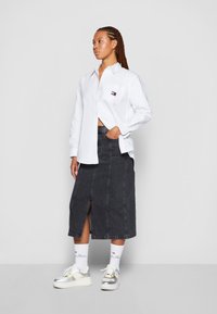 Biała koszula na guziki noszona na czarnej dżinsowej spódnicy z bocznym rozcięciem, w komplecie z białymi i srebrnymi sneakersami oraz skarpetkami z logo. Warkocze.