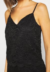 Camisole en dentelle noire avec bretelles spaghetti réglables, ornée de motifs floraux et d'un ourlet en forme d'ondulation. Texture douce et coupe ajustée.