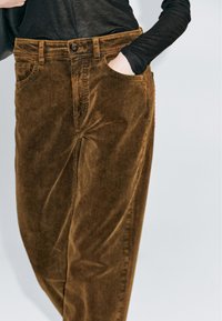 Bruine corduroy broek met een relaxte pasvorm, voorzien van zijzakken en een knoopsluiting bij de tailleband. Textuur met verticale ribbels zichtbaar.