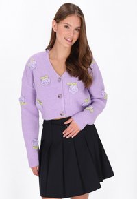 Fioletowy sweter robiony na druty z dekoltem w serek, ozdobiony haftowanymi różami i zielonymi liśćmi, z zapięciem na guziki i krojem w stylu cropped. Czarna spódnica plisowana.