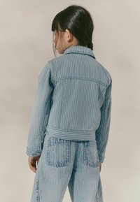 Lichtblauwe denimjasje met verticale witte strepen, kraag en verstelbare tailleband. Gestyled met bijpassende denim broek.